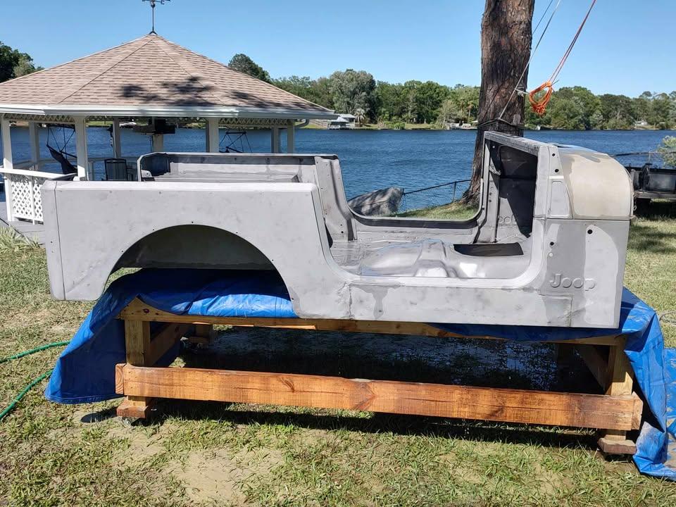 A gray, stripped-down Jeep body shell sits on a wooden platform outdoors near a lake. In the background, there is a gazebo, trees, and a blue sky. The Jeep has no wheels, doors, or interior parts.