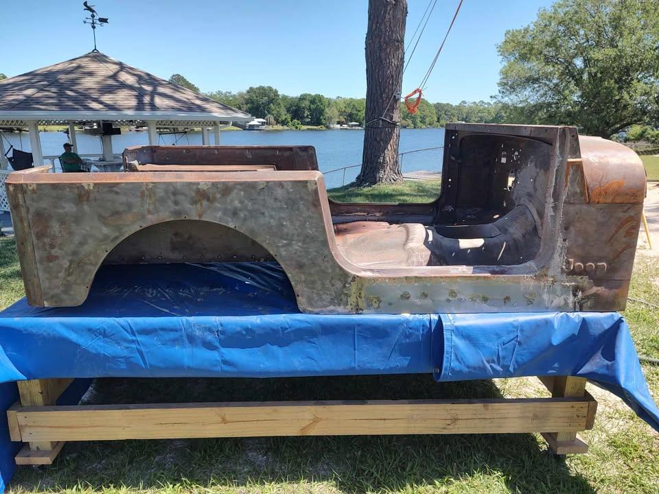 A rusty, stripped-down Jeep body sits on a wooden frame covered with a blue tarp outdoors, near a lake with trees and a gazebo in the background.
