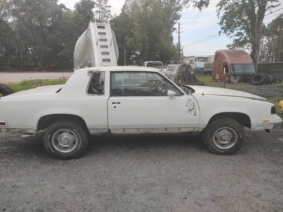 A white, older model two-door car with rust and visible damage is parked on gravel near industrial vehicles and trucks, with trees and power lines in the background.