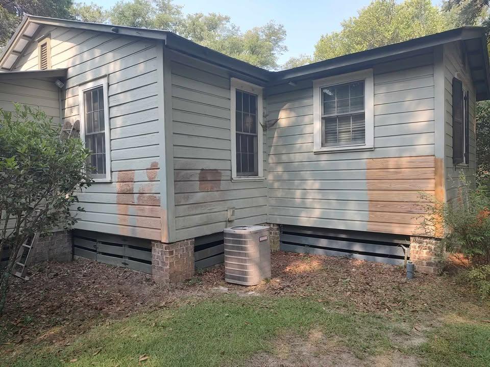 A light gray house exterior with several paint test patches in brown and tan shades on the siding. An air conditioning unit sits by the foundation, and there is some greenery and lawn in the foreground.