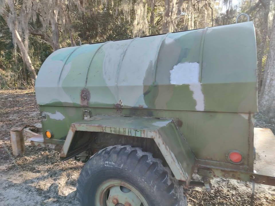 A green and white camouflaged utility trailer with a large cylindrical tank, parked outdoors on dirt near trees draped with Spanish moss. The trailer appears weathered and partially repainted.