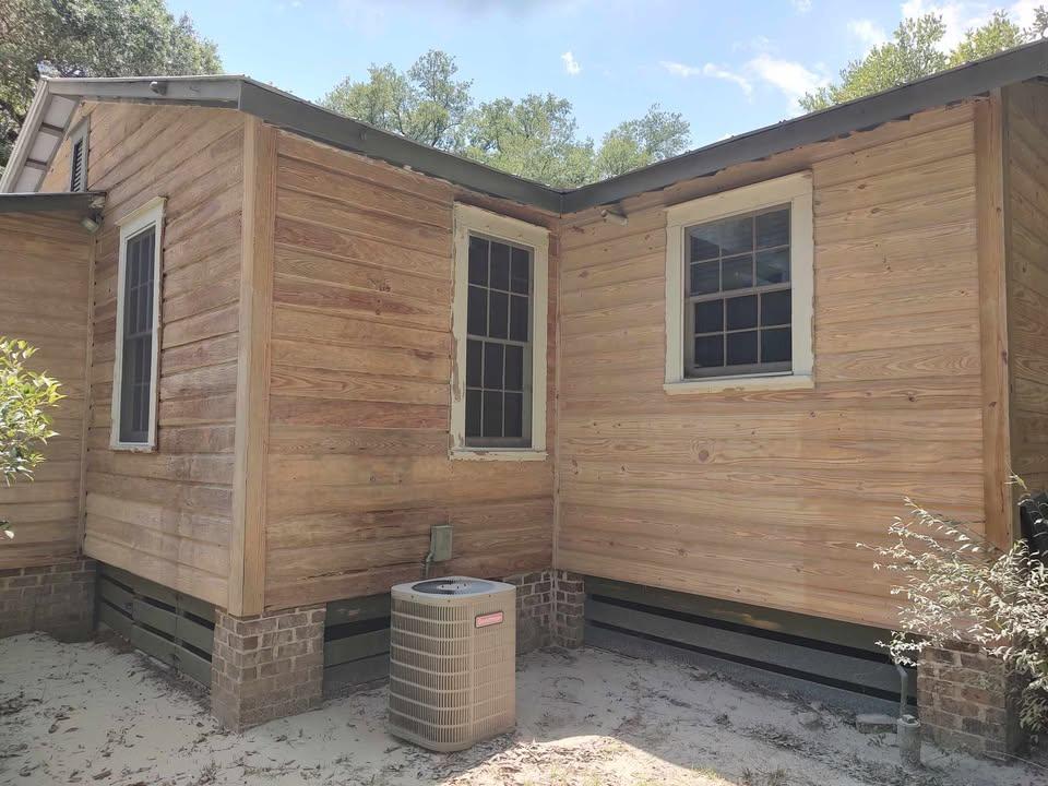 A wooden house exterior with two windows, light-colored wood siding, a sloped metal roof, and a central air conditioning unit on sandy ground beside the brick foundation. Trees and blue sky are in the background.