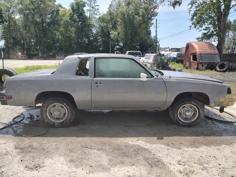 A gray two-door car with missing front and rear wheels is parked on a dirt lot, surrounded by trees and other vehicles in the background.