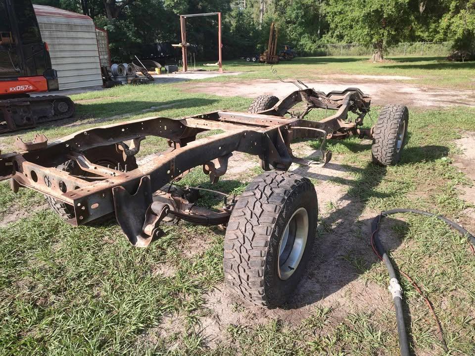 A rusty, stripped-down vehicle chassis with four tires sits on grass outdoors near a shed and construction equipment, with trees in the background.