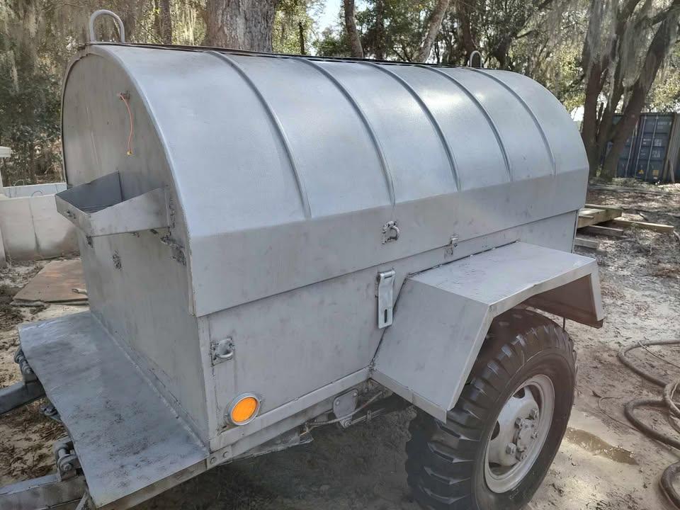 A silver, metal trailer with a rounded top and large tires is parked outdoors on dirt. The trailer has a rectangular compartment with a side door and latch, as well as a small orange reflector near the wheel. Trees are visible in the background.