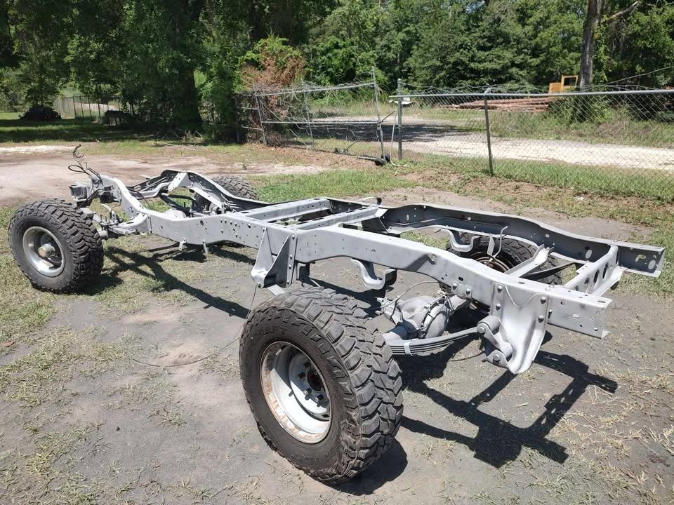 A metal vehicle chassis with large off-road tires is parked on dirt and grass outdoors, surrounded by trees and a chain-link fence in the background.