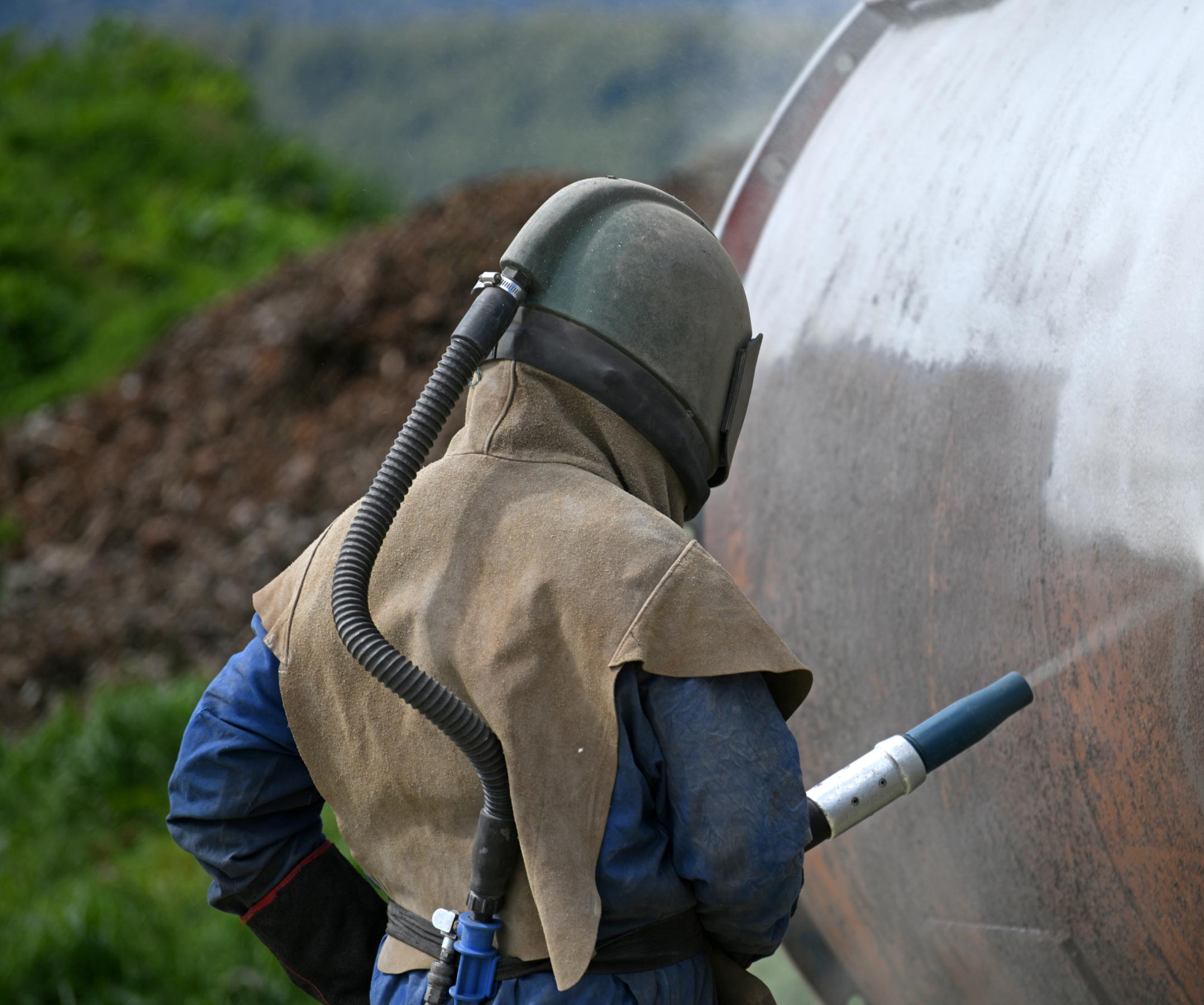 A person wearing protective gear and a helmet uses a sandblasting tool to clean or treat the surface of a large metal tank outdoors.