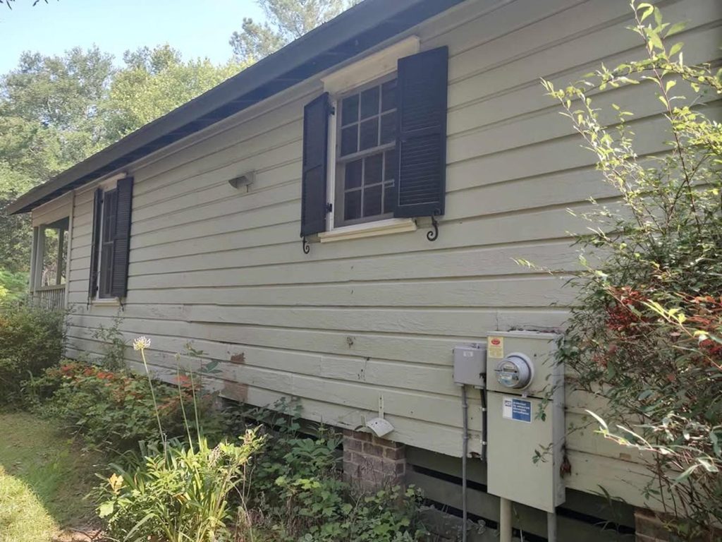 A light gray house exterior with several paint test patches in brown and tan shades on the siding. An air conditioning unit sits by the foundation, and there is some greenery and lawn in the foreground.