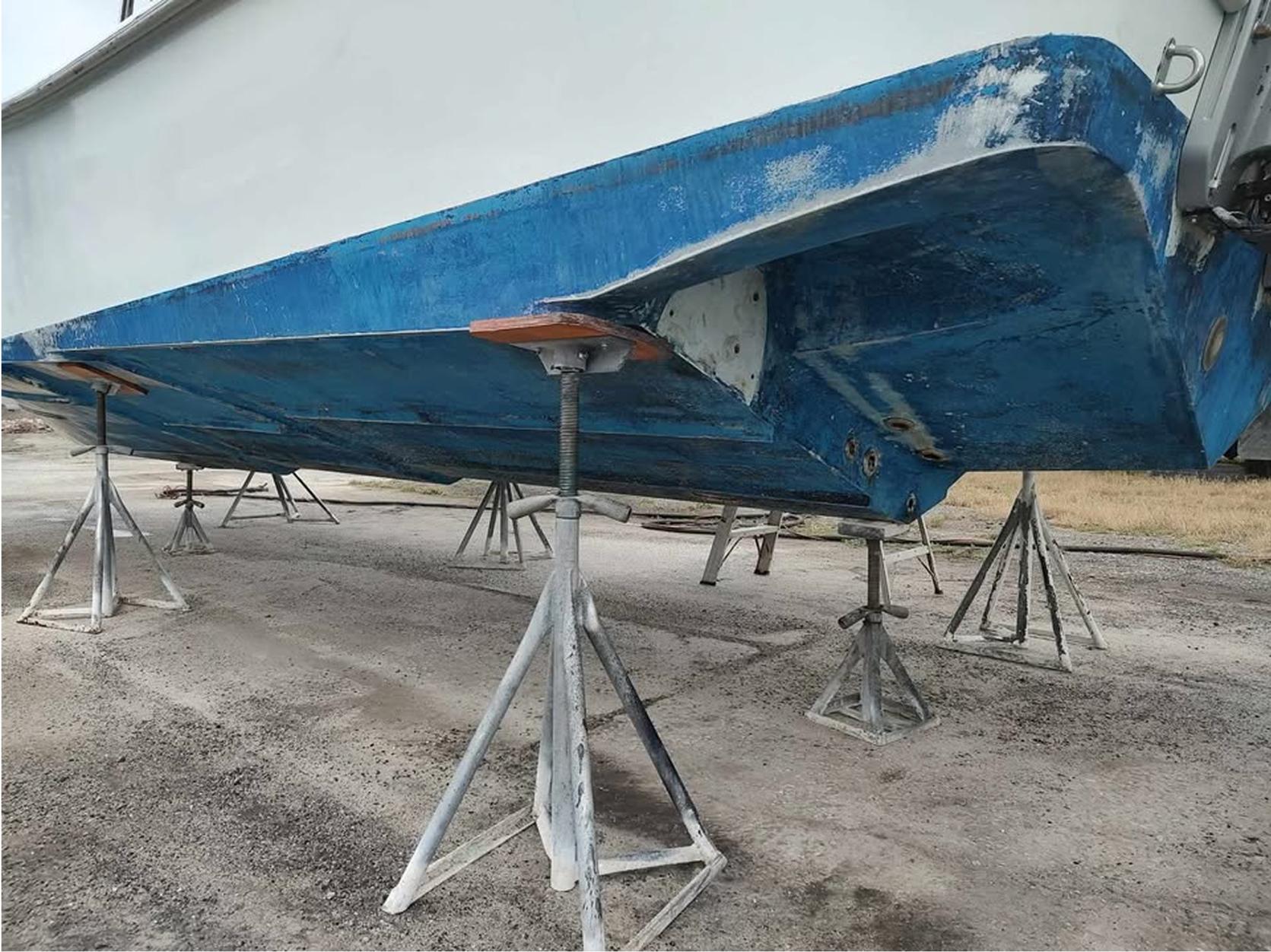 The underside of a boat with a blue hull is supported by multiple metal stands on a concrete surface, possibly in a boatyard for maintenance.