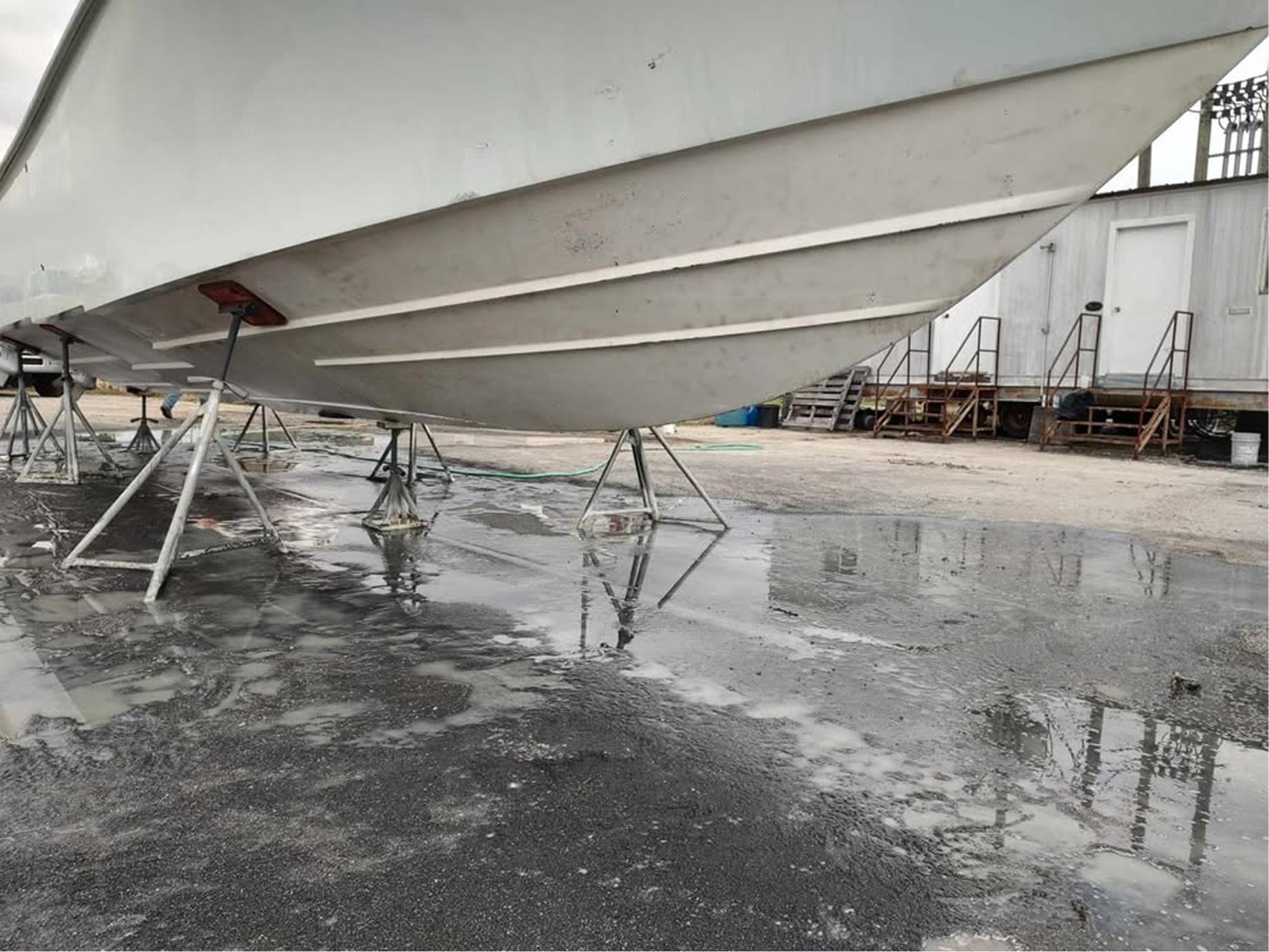 A large white boat is propped up on metal stands in a boatyard. The ground is wet with puddles reflecting the boats hull. There are small buildings and metal stairs in the background.