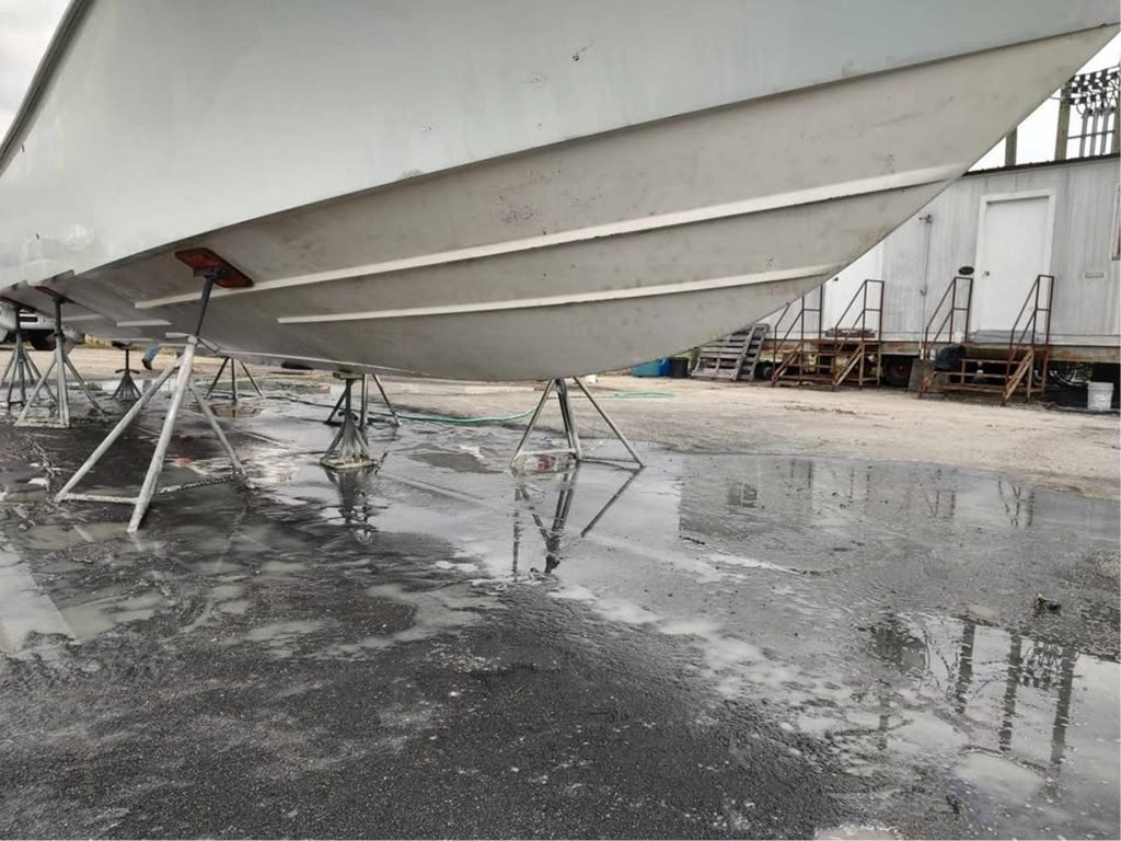 A large white boat is propped up on metal stands in a boatyard. The ground is wet with puddles reflecting the boats hull. There are small buildings and metal stairs in the background.