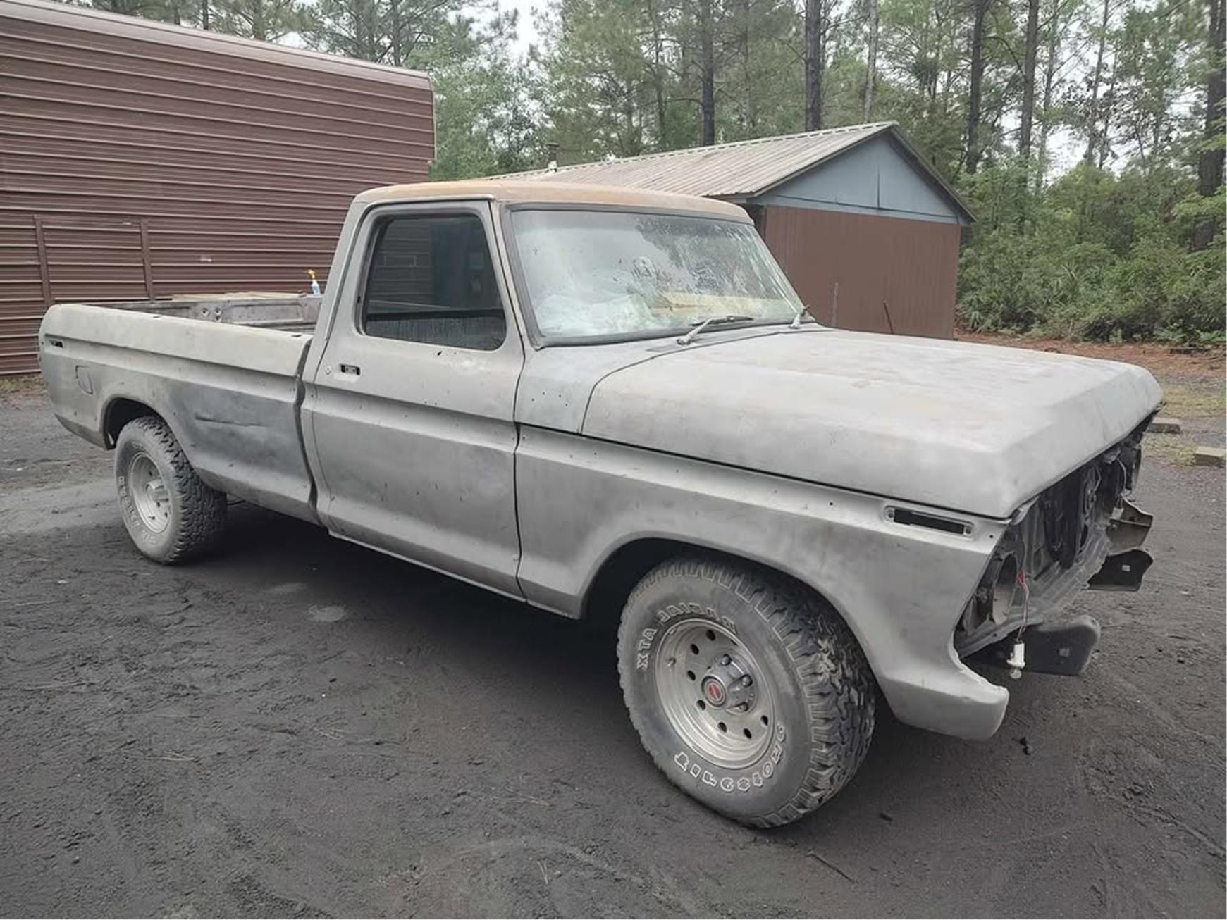 An old, weathered pickup truck without a front grille or headlights is parked on a dirt surface near a brown metal building and a shed, surrounded by trees.