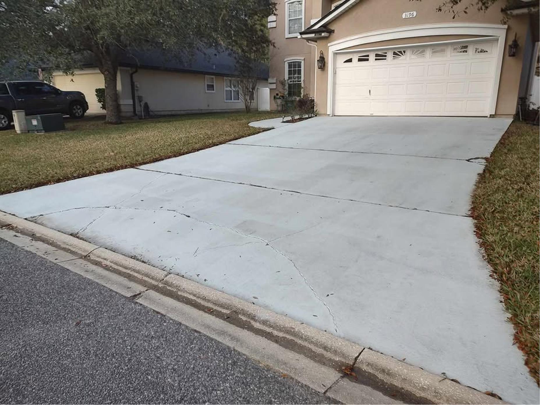 A light-colored concrete driveway with visible cracks leads to a double garage door on a beige house. Grass lawns border both sides, and a tree stands near the left side of the driveway.