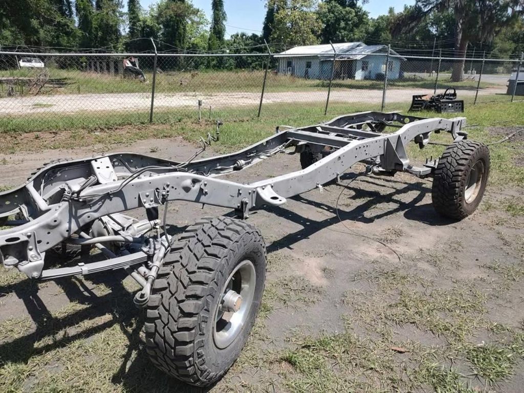 A metal vehicle chassis with large off-road tires is parked on dirt and grass outdoors, surrounded by trees and a chain-link fence in the background.