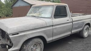 An old, weathered pickup truck without a front grille or headlights is parked on a dirt surface near a brown metal building and a shed, surrounded by trees.