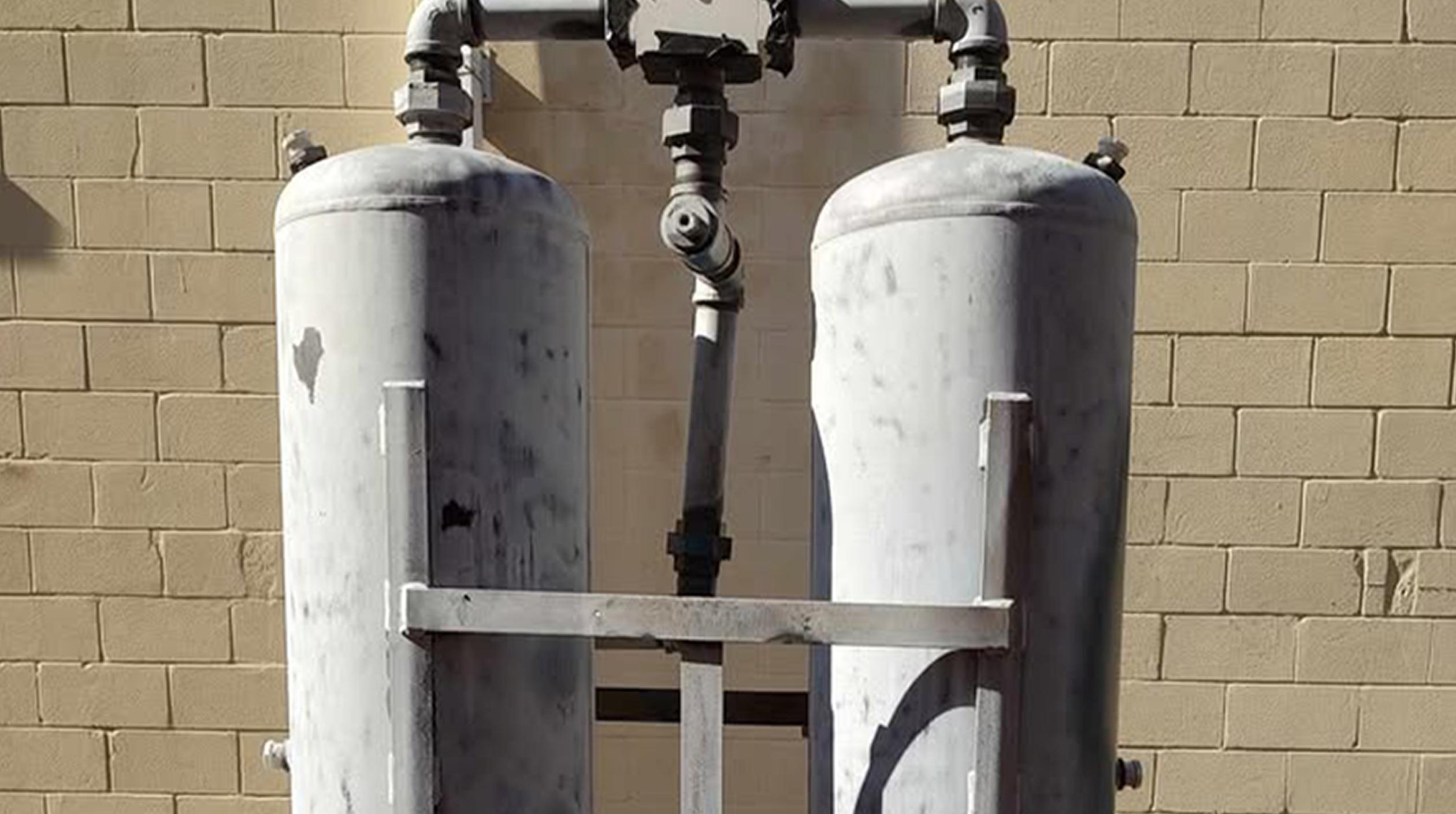 Two large, upright cylindrical metal tanks connected by pipes, positioned against a beige brick wall. The tanks are supported by a metal frame and appear weathered.