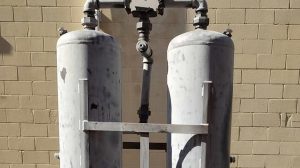 Two large, upright cylindrical metal tanks connected by pipes, positioned against a beige brick wall. The tanks are supported by a metal frame and appear weathered.