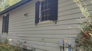 The side of a light-colored house with horizontal siding, a window with black shutters, electrical meter boxes mounted on the wall, and some plants growing nearby.
