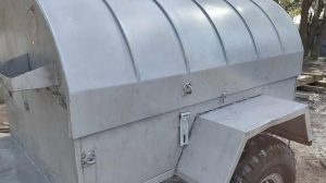 A close-up view of a metal trailer with a curved, domed top and large tires, parked outdoors on a dirt surface near trees. The trailer has a side compartment and a small orange reflector.