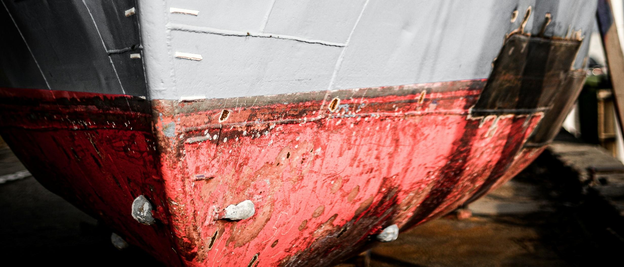 Close-up view of the weathered bow of a ship with chipped red and gray paint, rust stains, and visible metal bolts, positioned on dry land for maintenance or repair.
