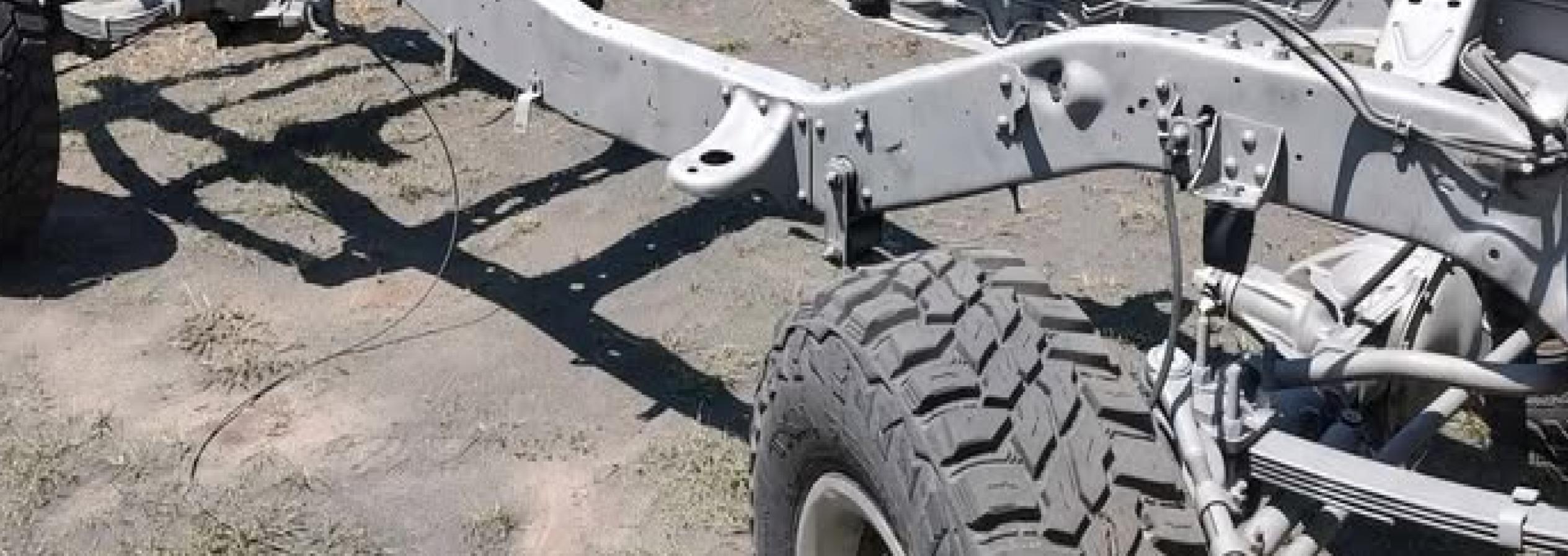 A metal vehicle chassis with large off-road tires is parked on dirt and grass outdoors, surrounded by trees and a chain-link fence in the background.