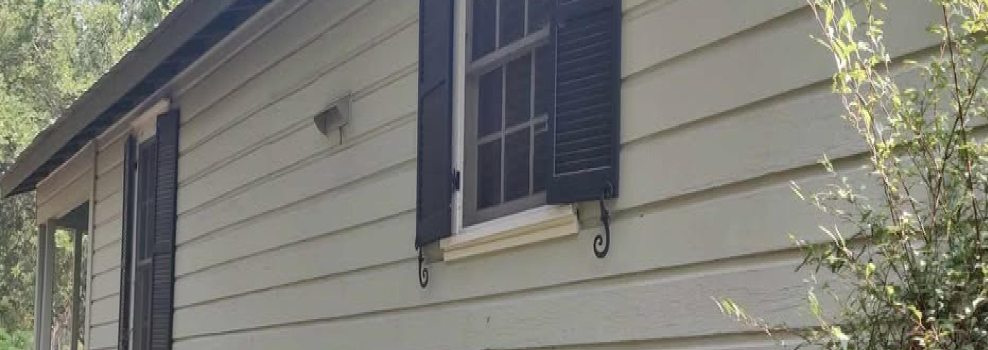 A light gray house exterior with several paint test patches in brown and tan shades on the siding. An air conditioning unit sits by the foundation, and there is some greenery and lawn in the foreground.