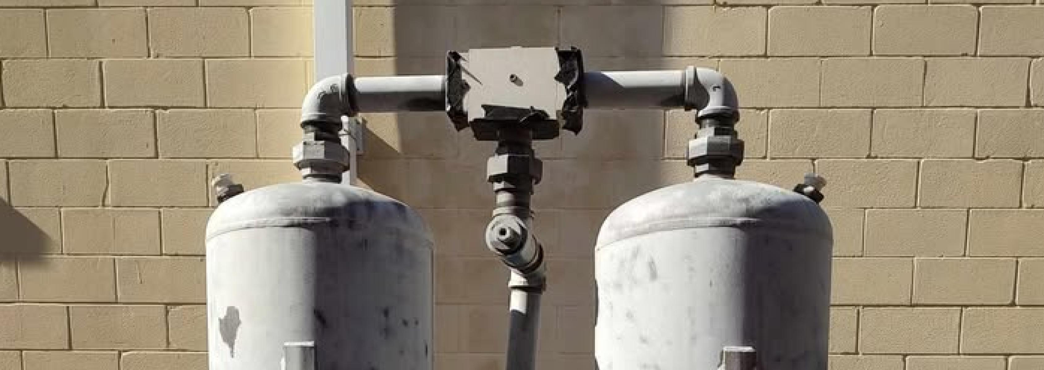 Two large, cylindrical metal tanks connected by pipes stand against a yellow brick wall, with visible bolts and fittings on the tanks and some discoloration on the surfaces.