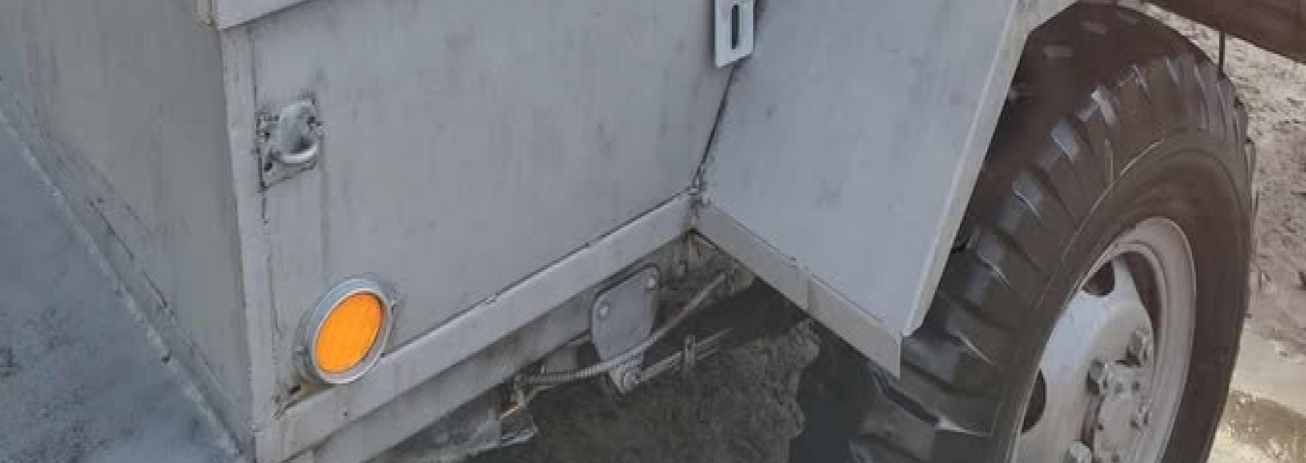 Close-up of the rear section of a gray utility vehicle, showing part of a large wheel, a yellow circular reflector, and metal panels with visible hinges and bolts.