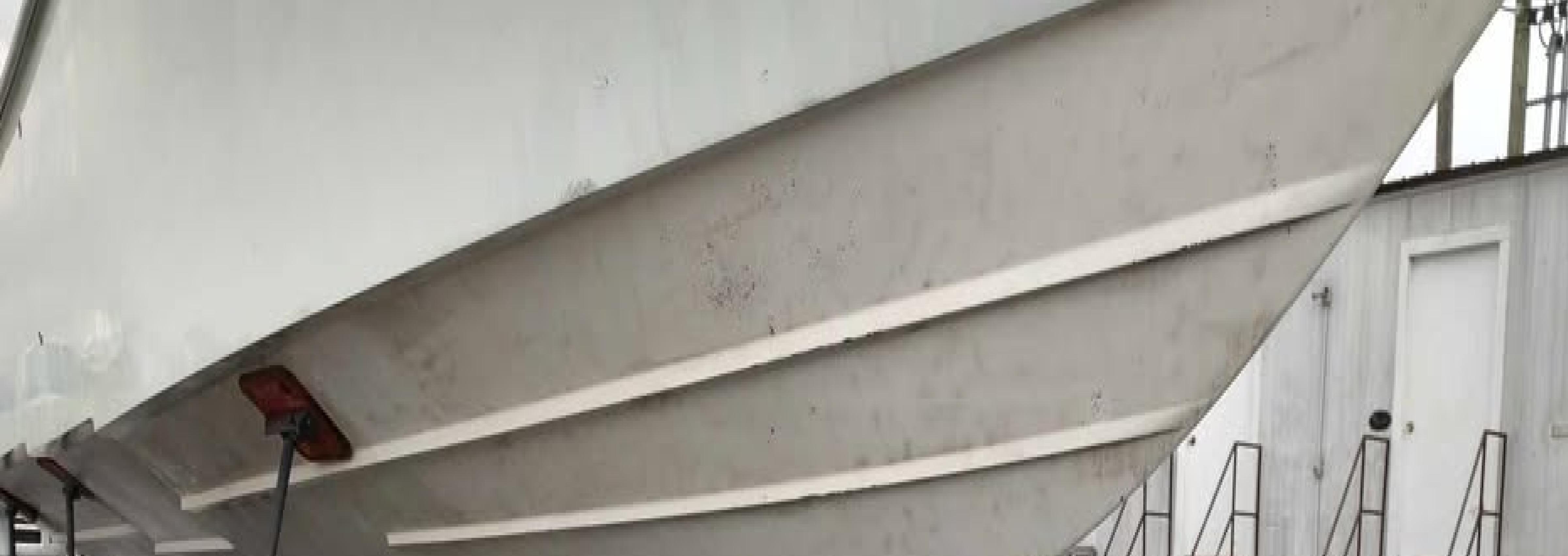 A large white boat is propped up on metal stands in a boatyard. The ground is wet with puddles reflecting the boats hull. There are small buildings and metal stairs in the background.