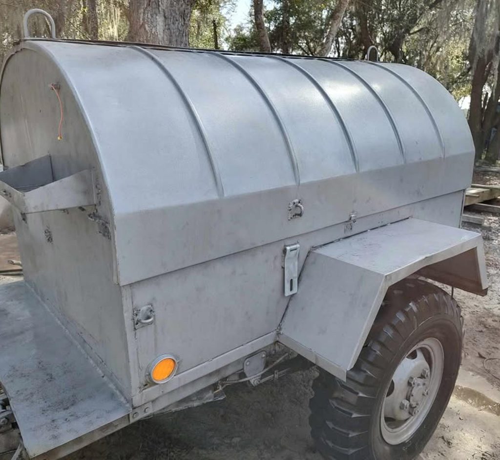 A close-up view of a metal trailer with a curved, domed top and large tires, parked outdoors on a dirt surface near trees. The trailer has a side compartment and a small orange reflector.