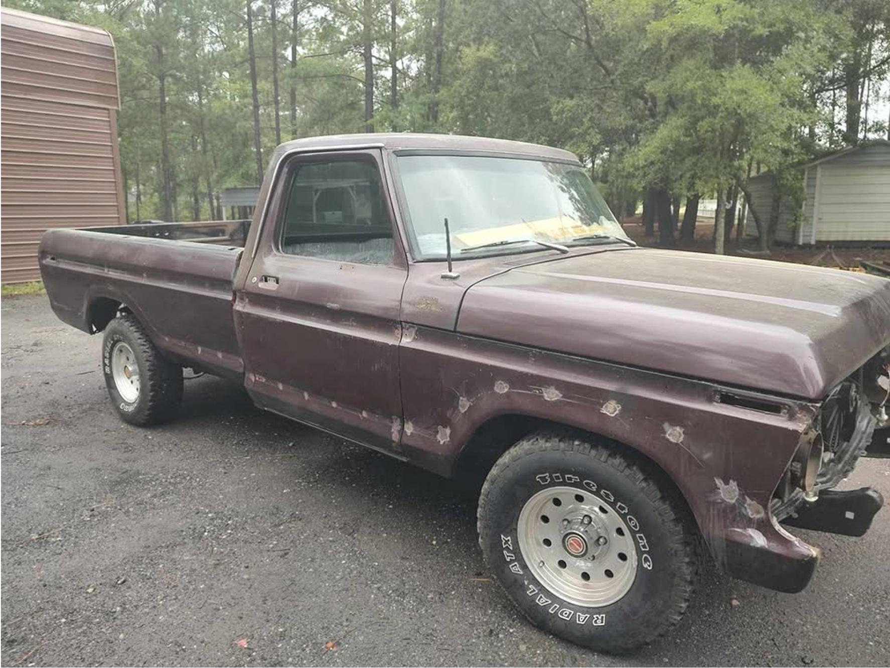 A vintage, purple pickup truck with visible dents and rust is parked on a gravel lot near trees and buildings. The truck’s front grill and headlights are missing, and the body shows signs of wear and repair.