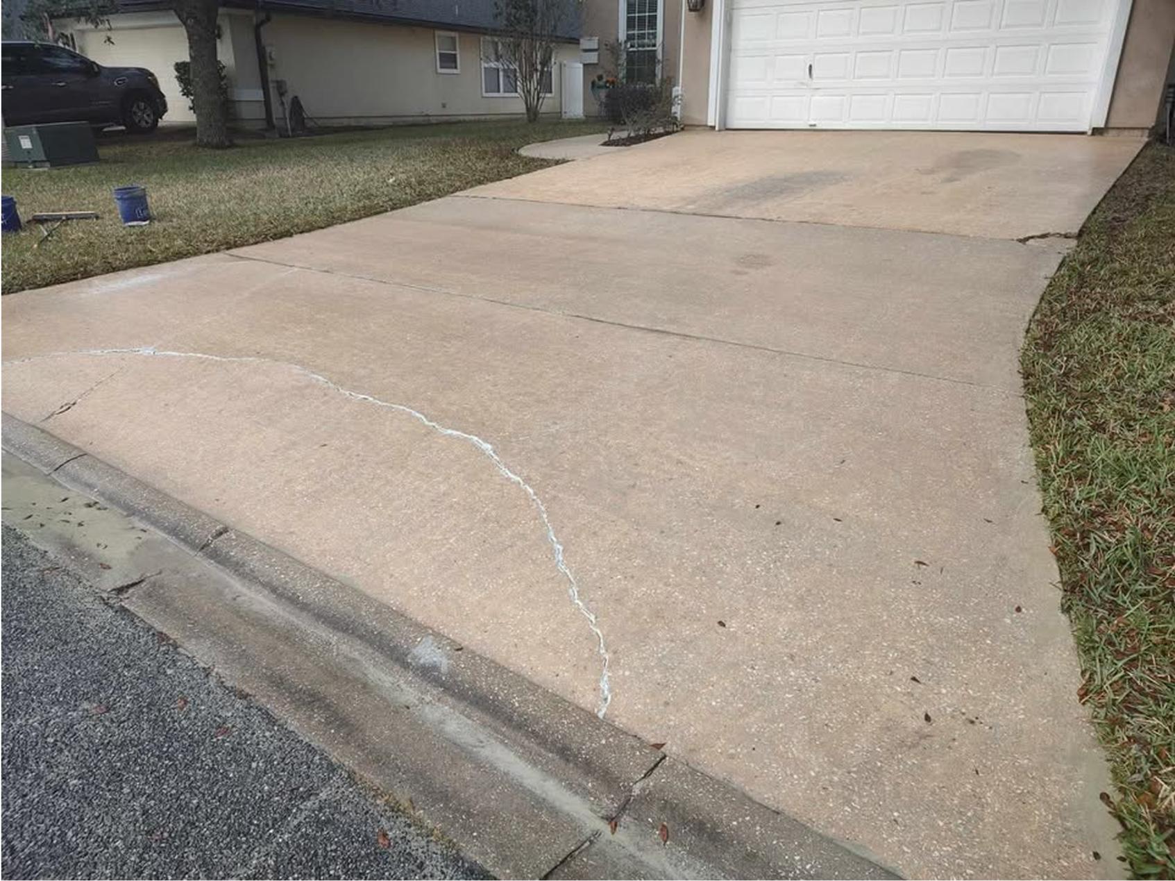 A light brown concrete driveway with a visible crack running across it, which has been filled with white sealant. The driveway leads up to a closed white garage door next to a grassy lawn.