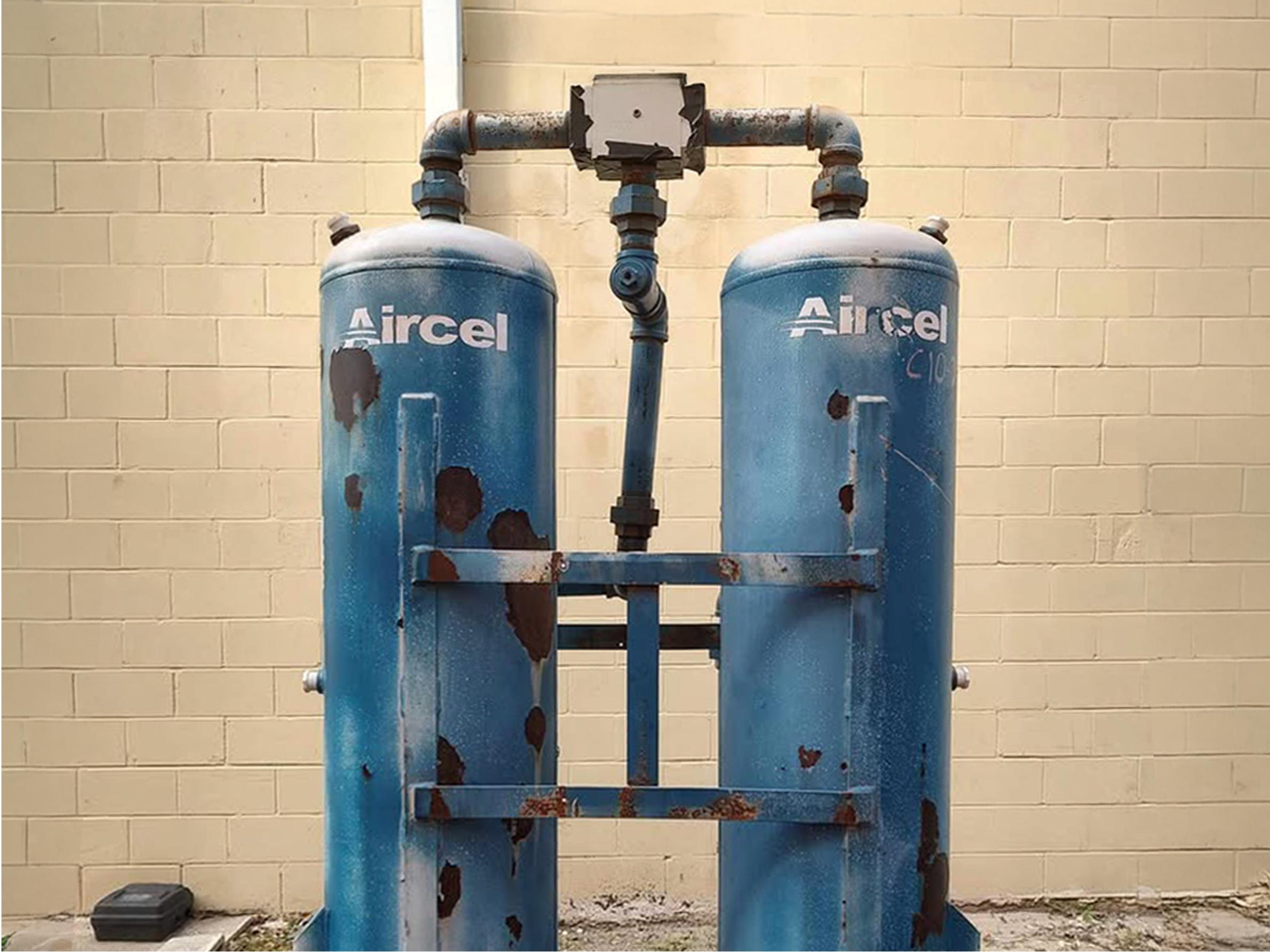 Two blue, rusted industrial tanks labeled Aircel stand side by side against a beige brick wall, connected by metal pipes at the top.